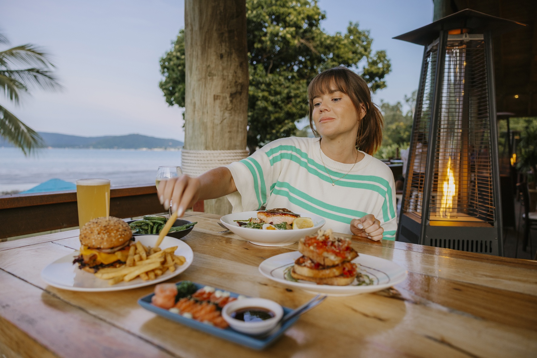 Rustic table with coffee, pastries, and citrus water on an outdoor terrace café at golden hour. Plush lounge chairs and wildflowers create a relaxing vibe. Panoramic views of green valleys and blue mountains glow in the soft, colorful sky. Dappled sunlight enhances the atmosphere.