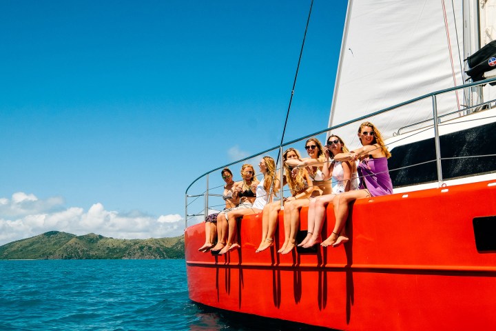 Seven female backpackers sitting on the side of a red sailboat with a blue sky and ocean backdrop.