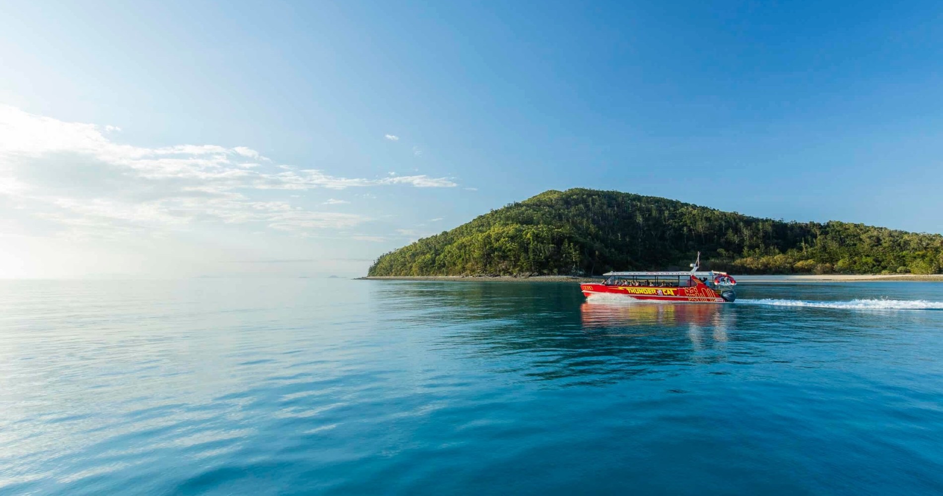 Modern tour boat cruises crystal-clear Whitsunday waters beneath midday sun, carrying diverse travelers enjoying ocean views, photography, and nature. Lush green islands, sparkling sea, coral shelf, and seabirds create a vibrant, adventurous Australian boat tour experience. Visible details include sunhats, sun-faded railings, and flying sea spray.