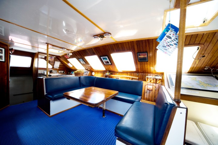 Spacious sailing boat interior with blue seating and wooden walls, featuring several windows and a central table.
