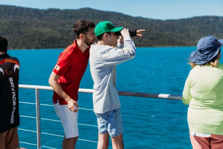 Staff member and guest standing on top deck, guest looking through binoculars with the ocean and mountains as backdrop