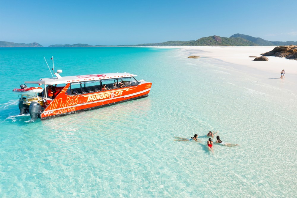 Small tour catamaran anchored at Whitehaven Beach, Australia, with sunbathers in the water getting ready to go snorkeling. Pristine white sand curves alongside turquoise water, while a few tourists walk near water’s edge.