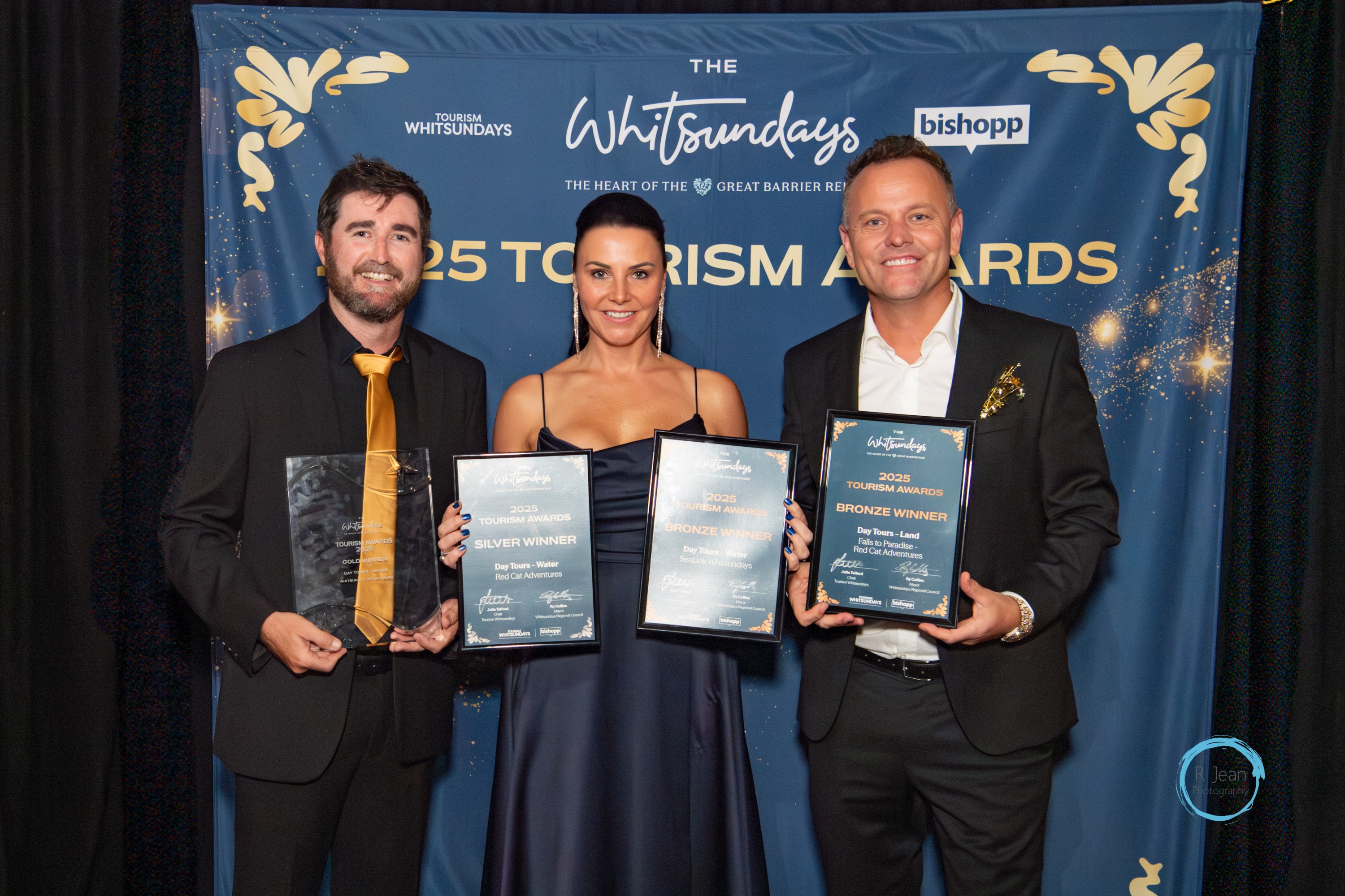 Three people holding awards at 2025 Tourism Awards with a blue and gold backdrop.