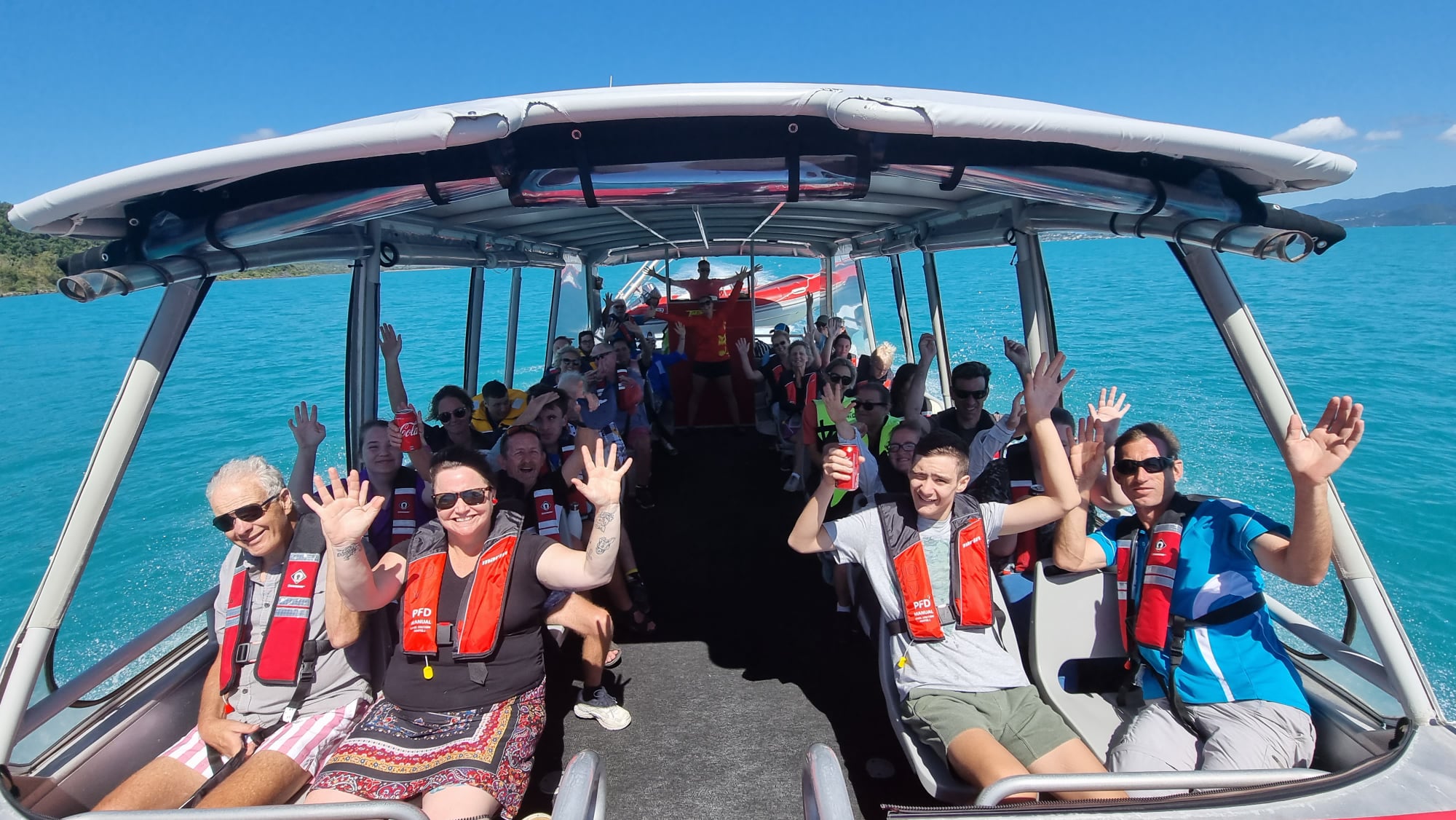 Diverse travelers aboard a sleek catamaran leaving Airlie Beach marina for a Whitsundays boat tour. Crew have assisted with lifejackets, tour boats underway, and lush green hills and palm trees frame the crystal-clear turquoise waters under a vibrant blue sky