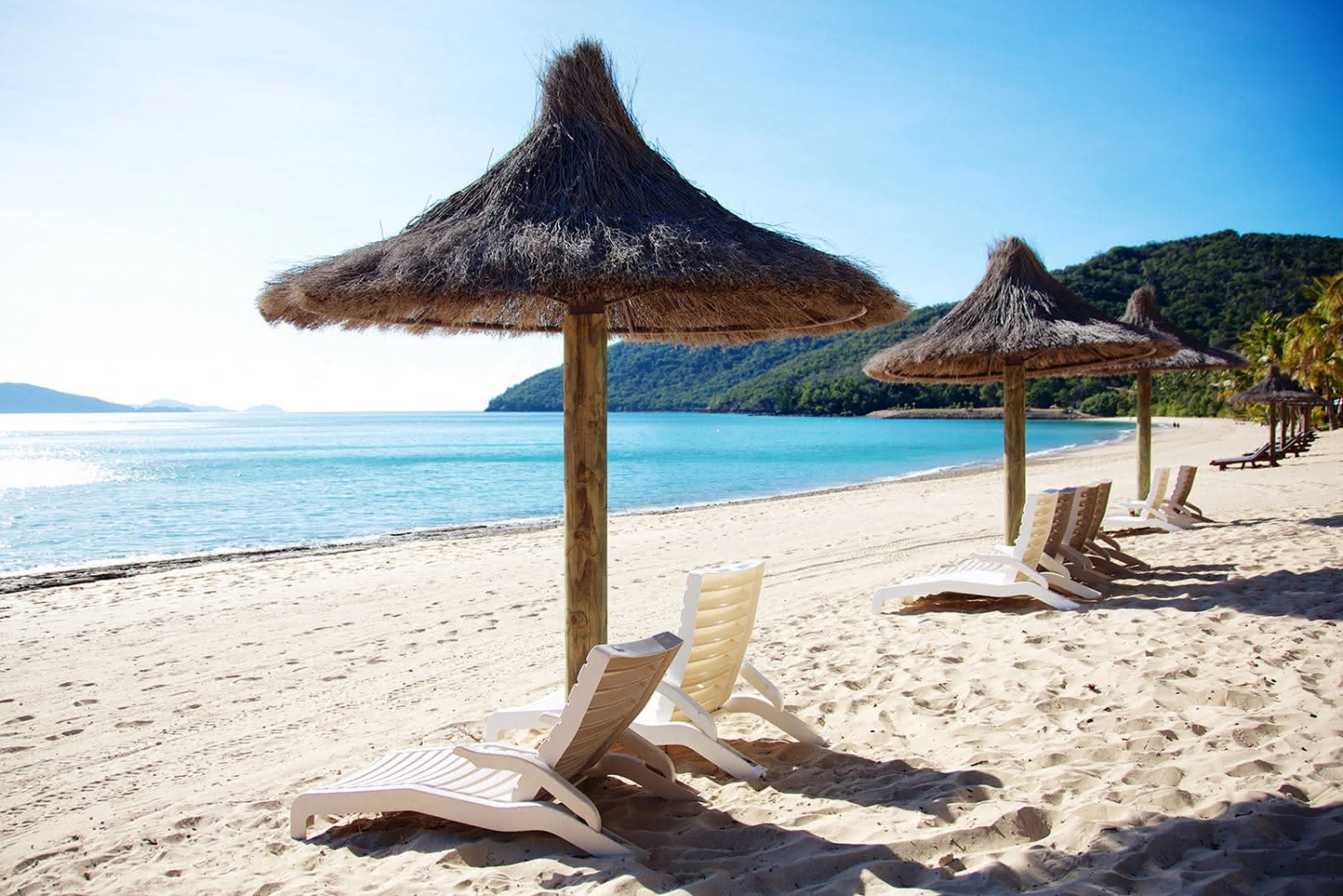 Wide-angle view of a serene beach with a tropical thatch umbrellas with sun loungers with palm tree lined tropical hills in the background all baked in golden morning sunlight.