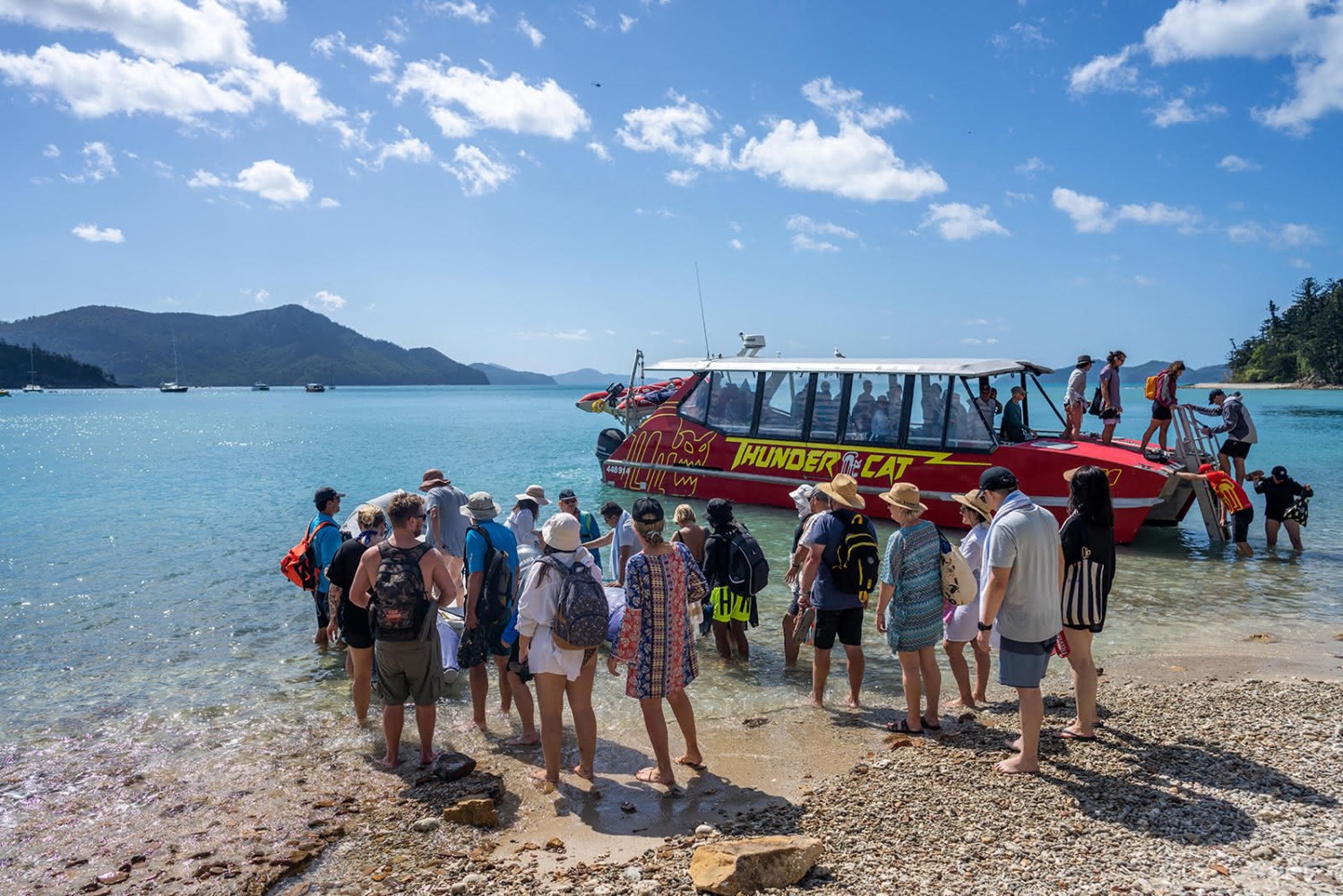A group of tourists disembark a luxury tour catamaran onto pristine Whitsundays beach, surrounded by turquoise water, snorkel gear, drinks, and a distant sandbar with eucalyptus trees, rainforest hills, and a few yachts in the background under a clear blue sky.