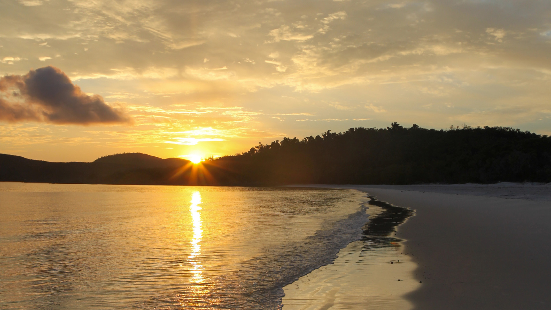 Wide-angle sunrise photo of Whitehaven Beach, showing swirling white silica sand, turquoise waves, rippled sand, seashells, footprints, and soft mist over distant islands.