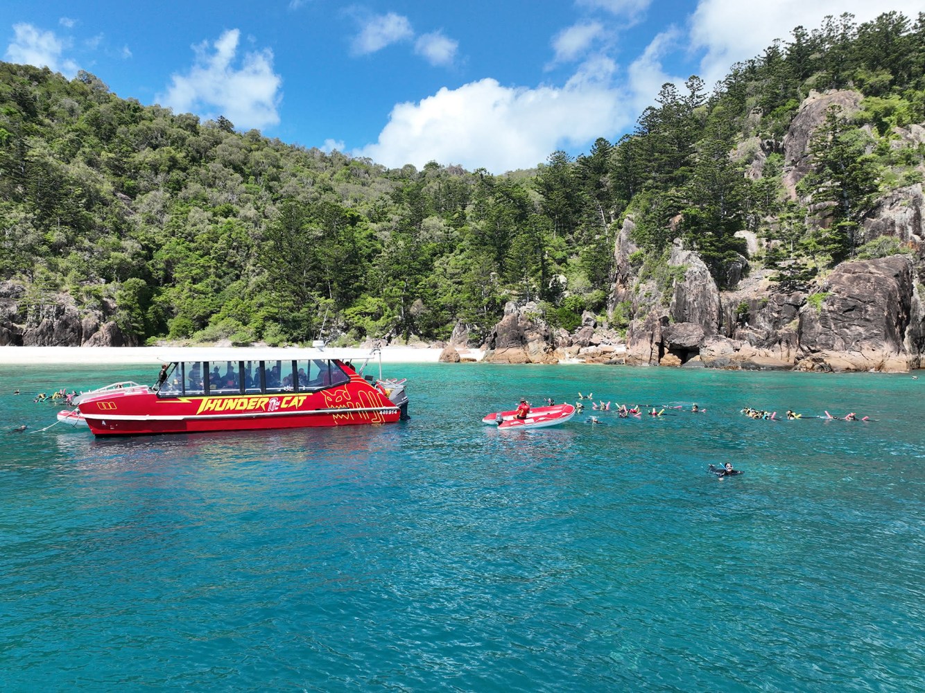 Red Cat Adventures tour boat in clear blue water near lush green cliffs and snorkelers enjoying teh corals reef below in aqua clear water.