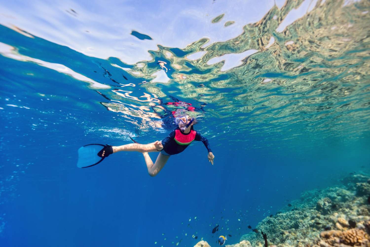 Person snorkeling underwater in clear blue ocean, wearing a snorkeling mask and fins near a coral reef.