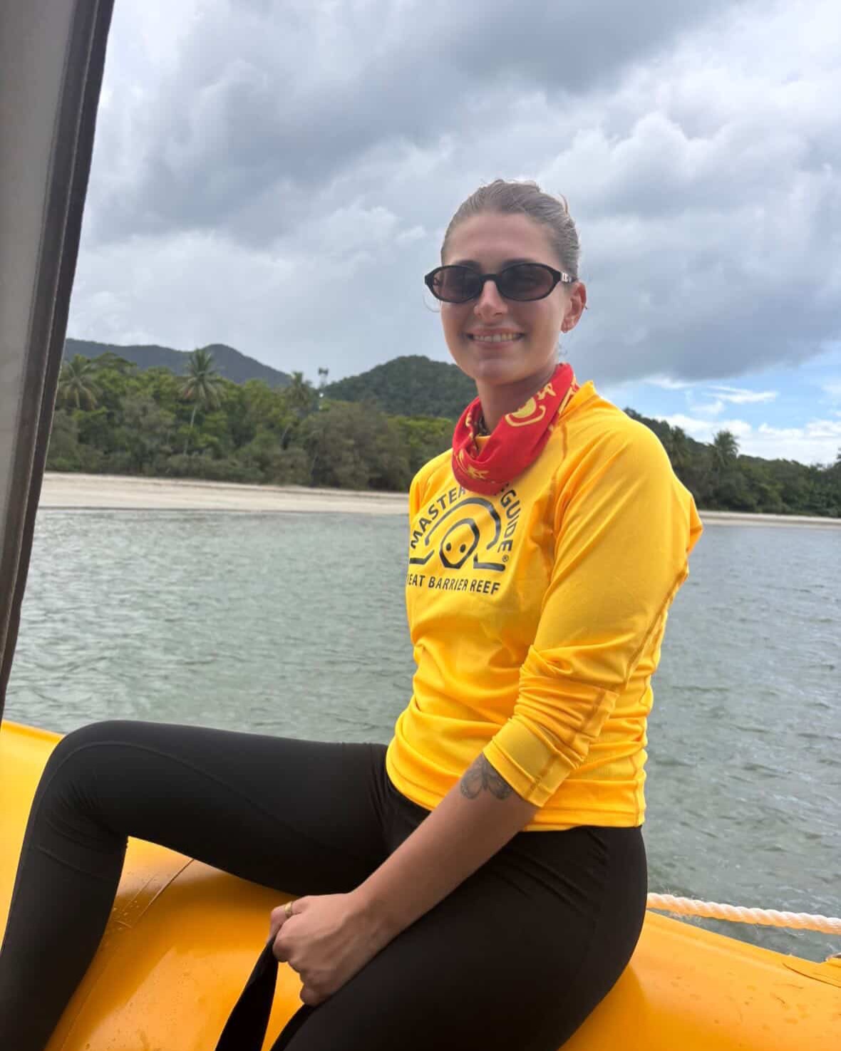 Person in yellow shirt and sunglasses sitting on a boat, with a beach and cloudy sky in the background.